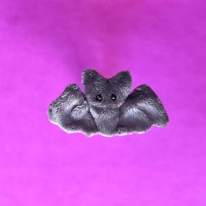 Pu&ograve; includere: Un giocattolo a forma di pipistrello grigio, con grandi ali spiegate e piccoli occhi neri. Il pipistrello &egrave; posto su uno sfondo magenta vibrante, creando un contrasto sorprendente. Il giocattolo ha una consistenza morbida e pelosa.