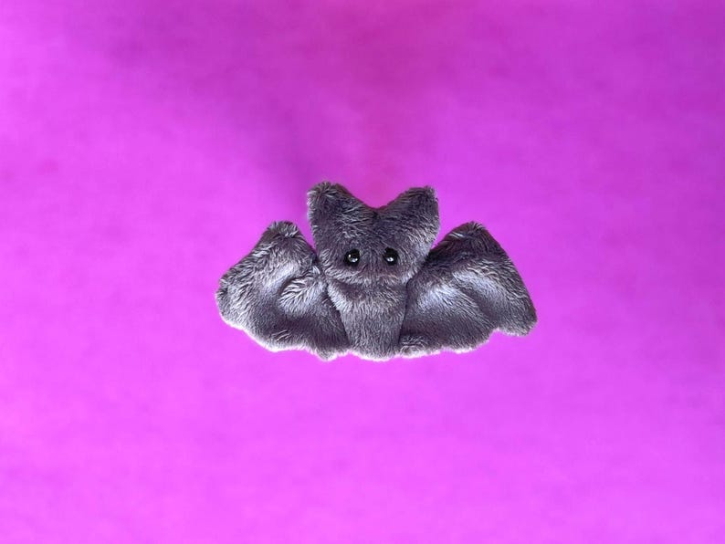 Pu&ograve; includere: Un giocattolo a forma di pipistrello grigio, con grandi ali spiegate e piccoli occhi neri. Il pipistrello &egrave; posto su uno sfondo magenta vibrante, creando un contrasto sorprendente. Il giocattolo ha una consistenza morbida e pelosa.