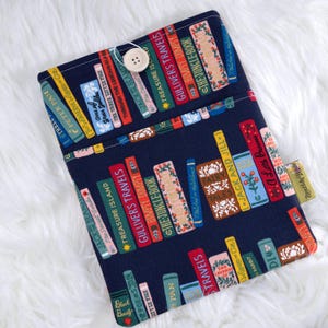 May include: A navy blue book sleeve featuring a colorful pattern of classic book spines. The spines display titles like "Gulliver's Travels" and "The Jungle Book." A wooden button secures the flap. The sleeve is designed to protect books.