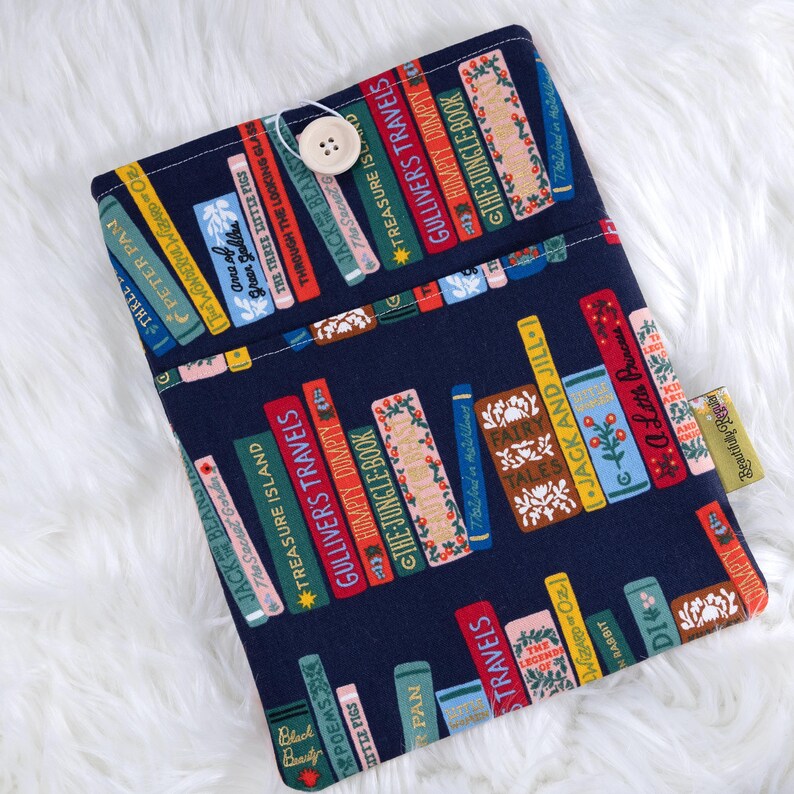 May include: A navy blue book sleeve featuring a colorful pattern of classic book spines. The spines display titles like "Gulliver's Travels" and "The Jungle Book." A wooden button secures the flap. The sleeve is designed to protect books.