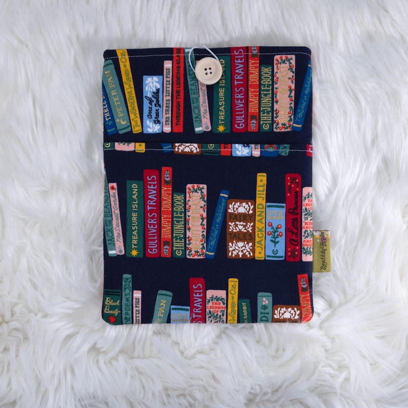 May include: A navy blue book sleeve featuring a colorful pattern of stacked books with titles like "Gulliver's Travels" and "The Jungle Book." The sleeve has a button closure and a small tag that reads "Beautifully." The background is a fluffy white surface.