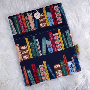 May include: A navy blue book sleeve featuring a pattern of colorful book spines. The spines display titles like "Gulliver's Travels," "Heidi," and "Jack and Jill." A wooden button and loop closure secures the sleeve. A small tag with text is visible.
