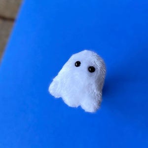 Puede incluir: Un peque&ntilde;o fantasma de peluche blanco con ojos negros de bot&oacute;n. El fantasma est&aacute; hecho de un material suave y difuso y est&aacute; sobre un fondo azul brillante. Este art&iacute;culo hecho a mano es perfecto para Halloween o como decoraci&oacute;n espeluznante.