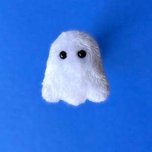 Puede incluir: Un peque&ntilde;o fantasma blanco de peluche con dos ojos negros de bot&oacute;n. El fantasma tiene una textura suave y difusa y una forma simple y redondeada. El fondo es azul brillante s&oacute;lido.