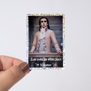 May include: A rectangular sticker with a holographic border. The sticker features a portrait of a person in a pink coat with the text "I can make the whole place Shimmer" in white. The person is standing on a balcony.