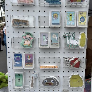 May include: A display of stickers and tarot cards in clear plastic holders on a white pegboard. The stickers feature various designs, including text, cartoon characters, and illustrations. Some cards are visible with illustrations and text.