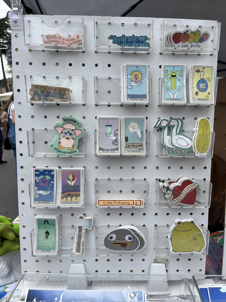 May include: A display of stickers and tarot cards in clear plastic holders on a white pegboard. The stickers feature various designs, including text, cartoon characters, and illustrations. Some cards are visible with illustrations and text.