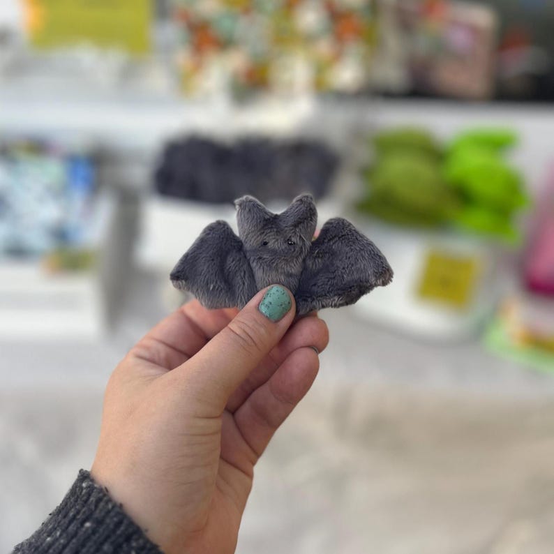 Pu&ograve; includere: Un piccolo pipistrello grigio di peluche con le ali spiegate. Il pipistrello ha un viso e orecchie dettagliati e un accento rosa sul petto. Il pipistrello &egrave; tenuto in una mano con un'unghia dipinta di verde acqua. Lo sfondo &egrave; sfocato.