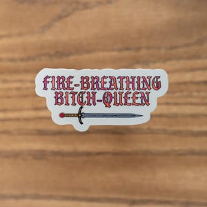 Op de afbeelding: Een witte sticker met de tekst "FIRE-BREATHING BITCH-QUEEN" in roze en oranje. Een zwaardillustratie staat onder de tekst. De sticker ligt op een houten oppervlak.