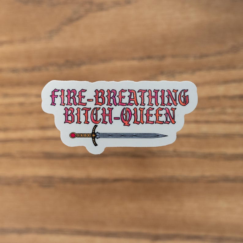 Op de afbeelding: Een witte sticker met de tekst "FIRE-BREATHING BITCH-QUEEN" in roze en oranje. Een zwaardillustratie staat onder de tekst. De sticker ligt op een houten oppervlak.