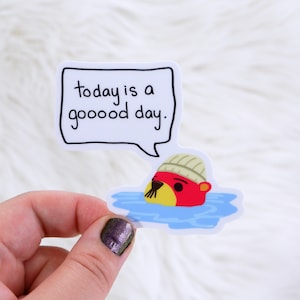 Peut inclure: Un autocollant représentant un phoque de dessin animé avec une tête rouge et un museau jaune portant un bonnet en tricot, nageant dans de l'eau bleue. Une bulle de dialogue au-dessus du phoque dit "today is a goooood day."