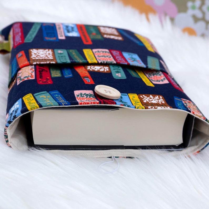 May include: A navy blue book sleeve with a colorful book print. The sleeve is secured with a wooden button. The interior is a light cream color. The book sleeve is designed to protect books.