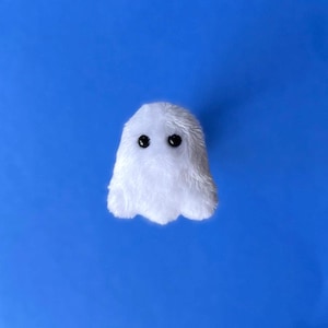 May include: A small, white plush ghost with two black button eyes. The ghost has a fluffy texture and a simple, rounded shape. The background is a solid, bright blue.