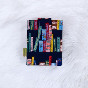 Puede incluir: Una funda de libro azul marino con una impresi&oacute;n colorida de libros apilados con t&iacute;tulos como "Peter Pan" y "Gulliver's Travels". La funda tiene un cierre de bot&oacute;n y una peque&ntilde;a etiqueta. El fondo es una superficie blanca y esponjosa.