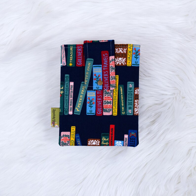 Puede incluir: Una funda de libro azul marino con una impresi&oacute;n colorida de libros apilados con t&iacute;tulos como "Peter Pan" y "Gulliver's Travels". La funda tiene un cierre de bot&oacute;n y una peque&ntilde;a etiqueta. El fondo es una superficie blanca y esponjosa.