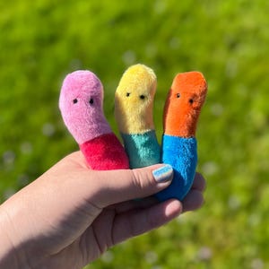 May include: Three colorful plush toys in pink, yellow, and orange, each with a contrasting bottom color. The pink toy has a red base, the yellow has a teal base, and the orange has a blue base. The toys have small black eyes.