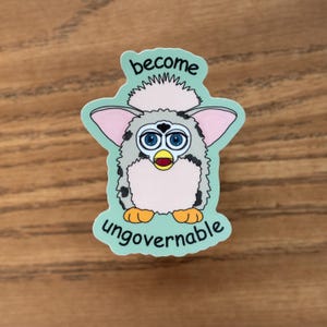 Puede incluir: Una pegatina con un personaje de dibujos animados parecido a un Furby, con ojos azules, orejas rosas y pies naranjas. El texto "become ungovernable" está encima y debajo del personaje sobre un fondo verde claro.