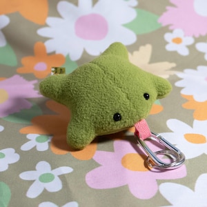 May include: A green plush toy in the shape of a star with black button eyes. It has a pink fabric loop attached to a silver carabiner clip. The background features a floral pattern with pink, white, and orange flowers.