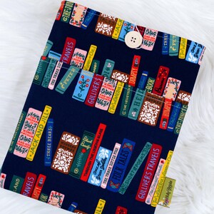 May include: A navy blue book sleeve featuring a colorful pattern of stacked books with titles like "Gulliver's Travels" and "Jack and Jill." The sleeve has a button closure and a small tag that reads "Beautifully Regular."