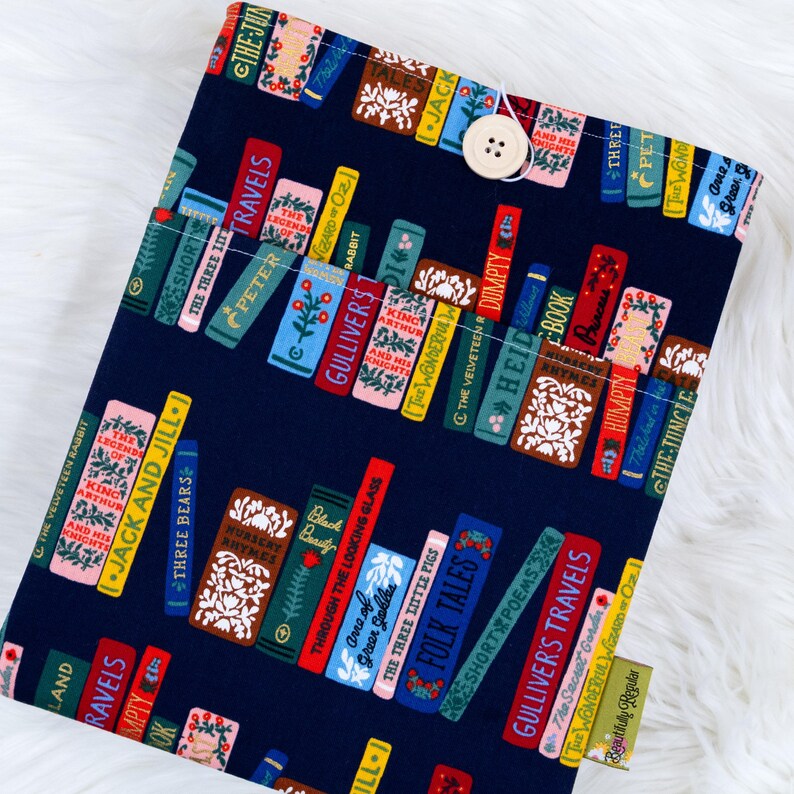 May include: A navy blue book sleeve featuring a colorful pattern of stacked books with titles like "Gulliver's Travels" and "Jack and Jill." The sleeve has a button closure and a small tag that reads "Beautifully Regular."
