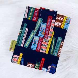 Puede incluir: Una funda de libro azul marino con un estampado de lomos de libros coloridos con t&iacute;tulos como "Gulliver's Travels" y "Peter Pan". La funda tiene un cierre de bot&oacute;n a presi&oacute;n y una peque&ntilde;a etiqueta que dice "Beautiful Reads".