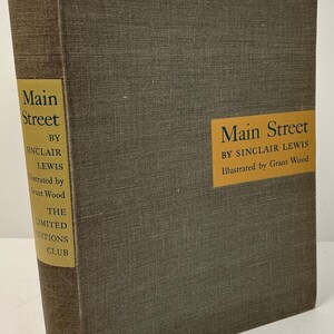 May include: A hardcover book titled "Main Street" by Sinclair Lewis, illustrated by Grant Wood. The book has a textured, gray-brown cover with gold lettering and a gold label. The Limited Editions Club is also printed on the spine.