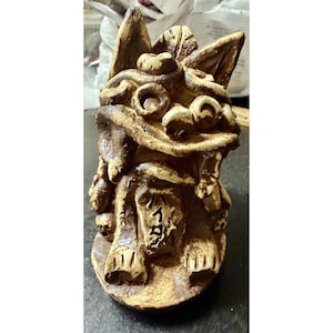 May include: A brown and cream-colored ceramic figurine of a mythical creature with pointed ears, swirling facial features, and a wide mouth. The figure has Japanese characters inscribed on its chest and sits on a circular base with a textured finish.
