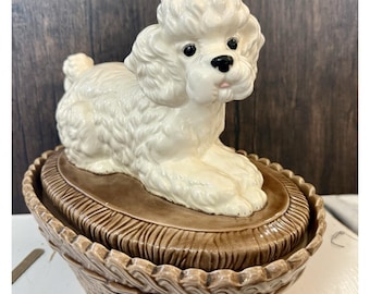 Tarro de cerámica vintage con forma de perro caniche en una cesta marrón, ideal como maceta o plato para dulces.