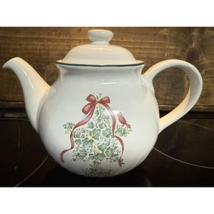 May include: White ceramic teapot with a lid and handle. The teapot is decorated with a design of green leaves, a red ribbon, and a small red bird. The rim of the lid and the top of the teapot have a green trim.