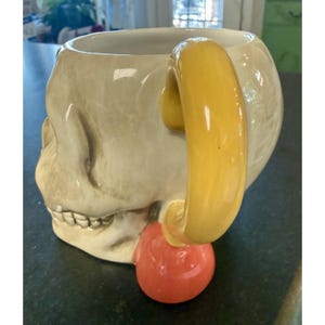 May include: A ceramic mug shaped like a human skull with a glossy off-white finish. The mug features a large, curved yellow handle attached to the side and a rounded, smooth orange base element at the bottom of the handle, near the jawline.