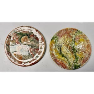 May include: Two circular ceramic coasters with textured surfaces. The left coaster features a white border surrounding a center with green, brown, and white streaks. The right coaster displays a mottled pattern of yellow, green, and brown tones.