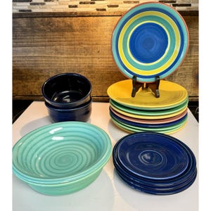 May include: A collection of ceramic dinnerware including a stack of multi-colored plates with concentric rings, a stack of solid dark blue plates, two dark blue bowls, and a single light green bowl with a spiral pattern on the interior surface.