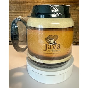 Peut inclure: Une grande tasse à café avec un couvercle et une poignée vert foncé, enveloppée dans du plastique transparent. La tasse a un design beige et marron avec les mots "Java Amore" et un logo de tasse à café. La tasse est posée sur une plateforme blanche.