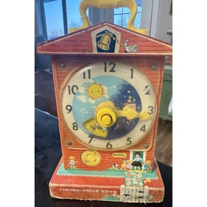 Op de afbeelding: Vintage Fisher-Price speelgoedklok in rood, met een gele handgreep. De wijzerplaat toont een dag- en nachtscène, met een gele knop in het midden. De klok heeft een schoolgebouwontwerp en de woorden "Fisher-Price Toys" staan onderaan.