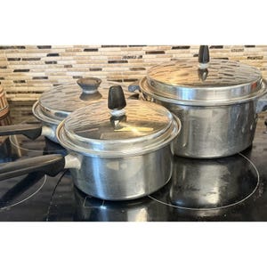 Op de afbeelding: Een set roestvrijstalen potten en pannen met glazen deksels en zwarte handgrepen. Het kookgerei staat op een zwart fornuis, met een patroon achterwand op de achtergrond. De potten variëren in grootte.