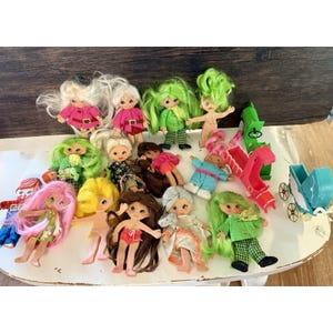 Könnte beinhalten: Eine Sammlung von Miniaturpuppen im Vintage-Stil mit verschiedenen Haarfarben und Outfits. Einige Puppen tragen rosa Mäntel, andere haben grüne, gelbe oder braune Haare. Zubehör: grüne Rutsche, blauer Kinderwagen und andere kleine Spielzeuge.