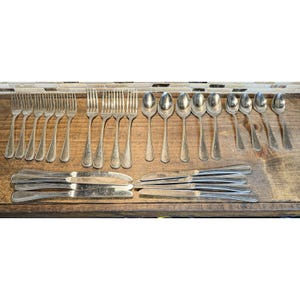 Puede incluir: Una colección de cubiertos antiguos, que incluye tenedores, cucharas y cuchillos, dispuestos sobre una superficie de madera. Los cubiertos son de metal brillante de color plateado. Los tenedores, cucharas y cuchillos están cuidadosamente organizados.