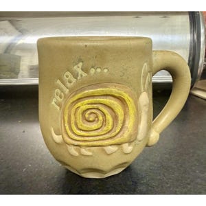 May include: A tan ceramic mug featuring a raised, embossed design of a snail with a yellow spiral shell. The word "relax..." is embossed in lowercase letters above the snail. The mug has a sturdy handle and a smooth, matte-textured finish.