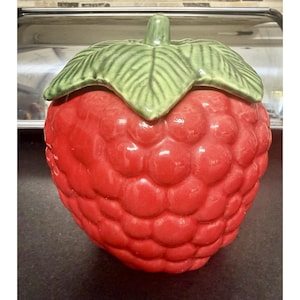May include: A ceramic jar shaped like a large red raspberry with a textured, bumpy surface. The lid is designed as a green stem with detailed leaves, featuring a central protrusion that acts as a handle for lifting the top of the container.