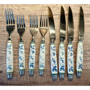 May include: A collection of vintage cutlery, comprising forks and knives. The handles are white with a blue floral pattern and green edging. The stainless steel blades and tines contrast with the decorative handles. The set is displayed on a wooden surface.