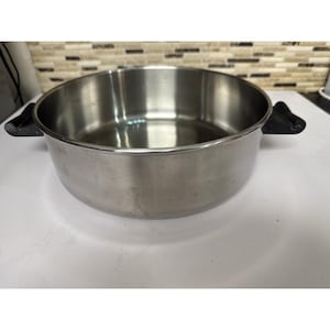 Puede incluir: Una olla de cocina de acero inoxidable con dos asas negras. La olla es redonda con una abertura ancha y un interior brillante. La olla está sobre una superficie blanca.
