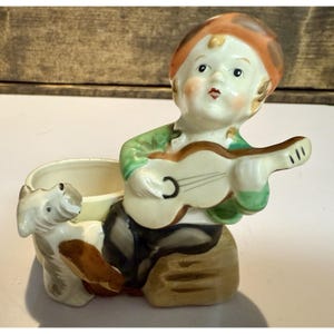 Könnte beinhalten: Ein Vintage-Keramik-Pflanzgefäß mit einer sitzenden Figur, die Gitarre spielt, mit einem Hund daneben. Die Figur trägt ein grünes Hemd, eine braune Hose und eine rote Mütze. Der Pflanztopf hat einen cremefarbenen Innenraum und ist detailliert bemalt.