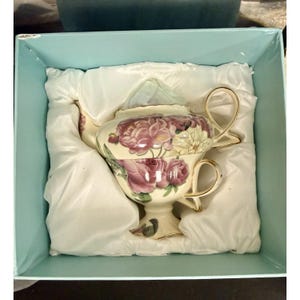 以下が含まれることがあります： ピンクと白のバラと緑の葉のフローラル柄が描かれたクリーム色の陶磁器製ティーポット。ティーポットの持ち手と注ぎ口には金色の縁取りが施されており、白いサテン生地が敷かれた水色の箱の中に収められています。