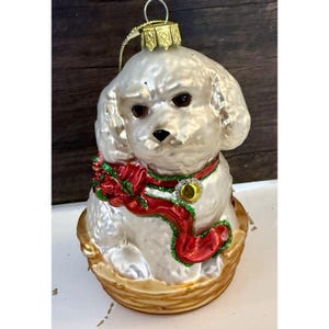 May include: A white dog Christmas ornament with a red ribbon and gold accents. The dog is sitting in a woven basket. The ornament has a gold cap and a gold string for hanging.