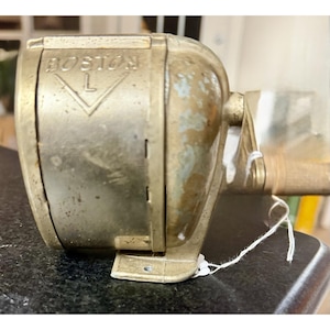 Puede incluir: Un sacapuntas manual vintage Boston L con un acabado metálico desgastado. El dispositivo presenta un logotipo en relieve que dice BOSTON L en la carcasa frontal, una manivela lateral y una base de montaje con un orificio para tornillo.