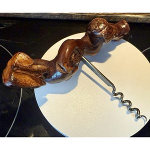 May include: A handcrafted wooden corkscrew with a natural, organic shape. The handle is made of polished, dark brown wood, and the metal corkscrew is silver. The corkscrew is resting on a white surface.