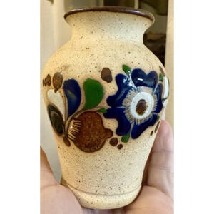May include: A small, speckled beige ceramic vase with a narrow neck and a flared rim. The surface is decorated with hand-painted floral motifs in deep blue, vibrant green, and earthy brown tones, featuring bold petals and leafy accents.