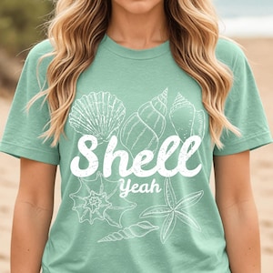 Puede incluir: Una camiseta verde agua con un diseño blanco de varias conchas marinas y una estrella de mar. La palabra "Shell" está escrita en negrita, cursiva, encima de la palabra "Yeah".