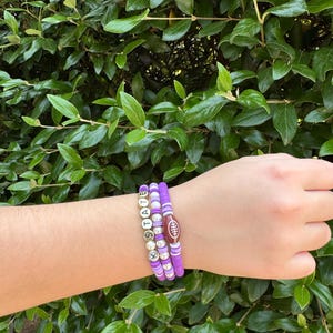 May include: Three purple beaded bracelets. One bracelet spells out "STATE" with silver letter beads. The second bracelet has a football-shaped bead. The third bracelet is made of purple beads. The bracelets are on a person's wrist.