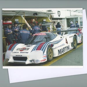 Könnte beinhalten: Ein Oldtimer-Rennwagen mit Martini-Lackierung, weiß mit roten und blauen Streifen, steht in der Boxengasse. Das Auto hat die Nummer 4. Personen in Rennteam-Uniformen sind im Hintergrund.